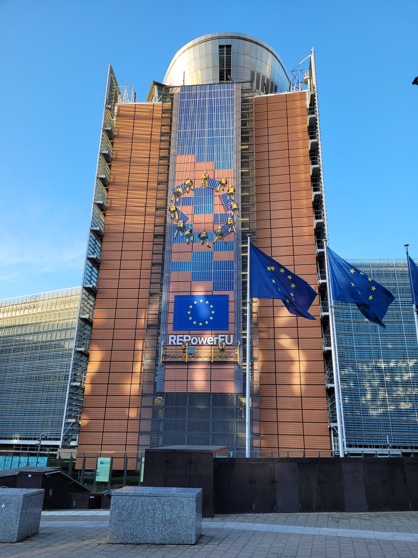 Turm des EU-Parlaments mit dem Sternenkreis im Zentrum, darunter die EU-Flagge und die Aufschrift "REPowerEU". Vor dem Gebäude stehen drei EU-Flaggen. 